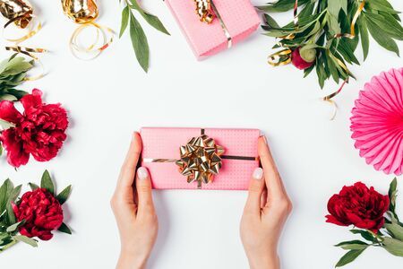 Woman's hands holding pink gift box with golden bow among lush peony flowers and holiday decorations on white background.の写真素材