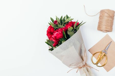 Festive flat lay composition bouquet of peony flowers packed in brown eco paper next to jute rope and golden scissors on white table with copy space.の写真素材