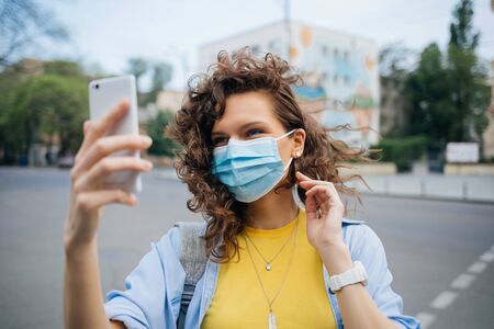 Happy young woman in medical mask for protection against the virus stands on city street makes selfie using mobile phone.の写真素材
