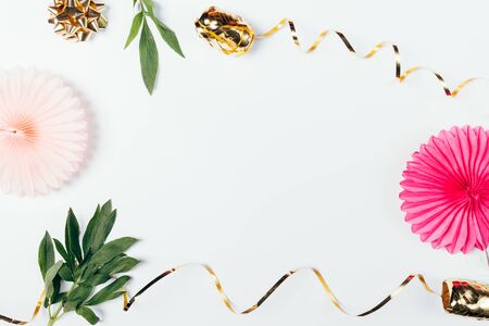 Festive summer flat lay frame of golden streamer ribbons and pink paper ornaments next to green leaves on white background with free space in center.の写真素材