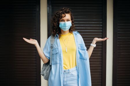 Closed stores during quarantine, young woman in protective medical mask stands on the threshold of non-working shop in confusion.の写真素材