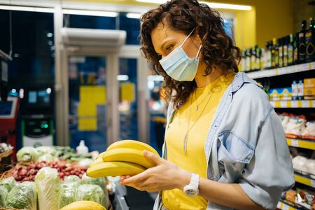 Young woman wearing medical protective mask choosing groceries while standing near fruit shelf in supermarket.の写真素材