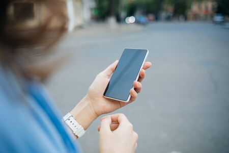 Mobile phone with blank screen in hand of young woman walking along city street in daytime, close-up.の写真素材