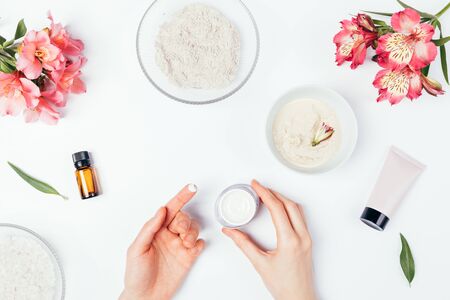 Flat lay composition woman's hands apply cosmetic cream next to ingredients for homemade clay facial mask, beauty skin care products and fresh flowers.の写真素材