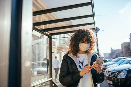 Young beautiful woman wearing medical face mask standing at bus stop in European city and holding her mobile phone.の写真素材