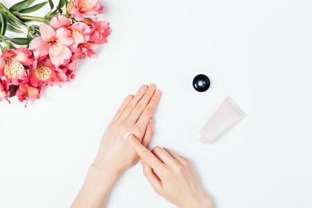 Woman's hands apply nourishing skin care cream next to bouquet of pink flowers on white table, top view.の写真素材
