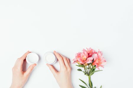 Female hands holding open jar of moisturizer cream next to delicate pink flowers on white background, flat lay.の写真素材