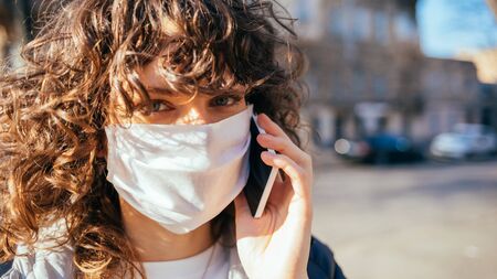 Young woman wearing protective face mask calling on mobile phone while standing on street in the city.の写真素材