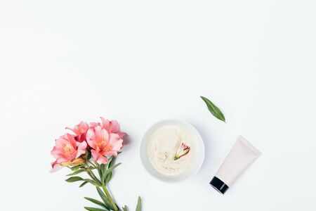 Cosmetic clay mask for skin care next to tube of cream and fresh flowers on white background with copy space.の写真素材