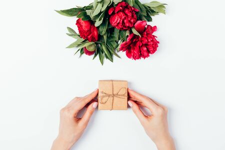 Top view female hands holding small brown gift box next to bouquet of lushly blooming peony flowers on white background.の写真素材