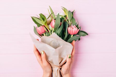 Top view of female hands holding lush bouquet of tulips in paper packaging on pink wooden table.の写真素材