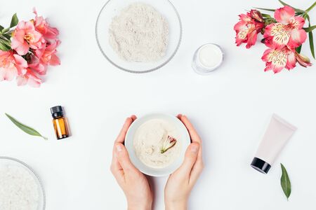 Woman's hands holding bowl homemade facial clay mask next to beauty skin care products cream, lotion and fresh flowers on white background, flat lay.の写真素材