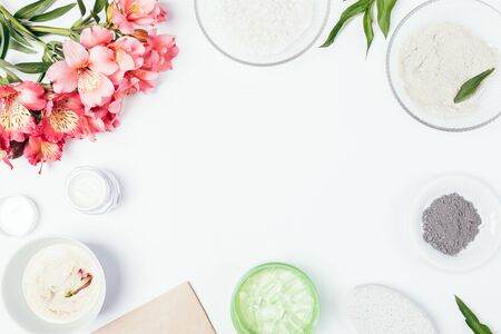 Flat lay frame of ingredients for homemade skin care cosmetics, creams and fresh flowers on white background with free space in center.の写真素材