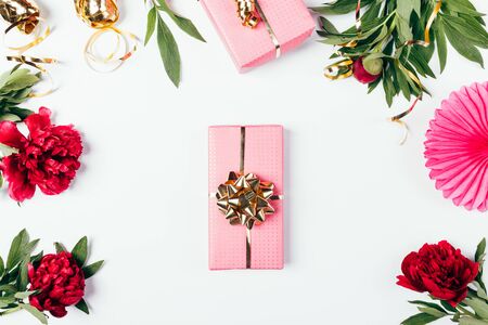 Pink gift box with golden bow among lush peony flowers and holiday decorations on white background.の写真素材