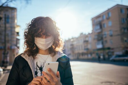 Portrait young woman wearing medical face mask as protection from the virus looking at her mobile phone, standing on city street, copy space.の写真素材