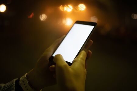 Close-up young woman hands typing in mobile phone with blank screen on illuminated city street at night.の写真素材