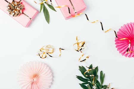 Pink gift boxes, paper birthday party decorations next to golden streamers and green leaves on white background, flat lay.の写真素材