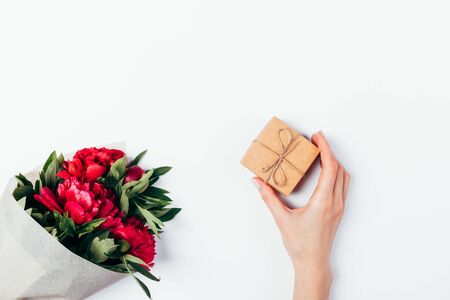 Female hand holds small gift box of craft paper tied with jute rope next to bouquet of fresh peony flowers on white background, flat lay.の写真素材
