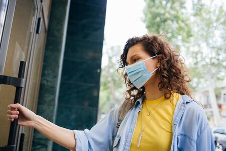 Portrait young woman wearing protective medical mask holds on to the handle and opens the door when entering store.の写真素材