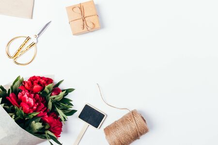Floral flat layout of bouquet of peonies wrapped in eco paper next to jute rope, gift box, scissors and wooden chalk plate on white background with copy space.の写真素材