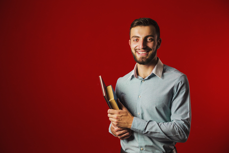 A portrait shot of a attractive handsome smiling student beard man with books on red backgroundの写真素材