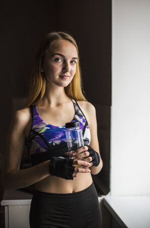 Attractive blonde girl in purple headphones with bottle in hand listening musci near the windowの写真素材