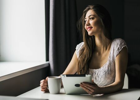 A attractive young brunette girl works with tablet pc in kitchen near windowの写真素材