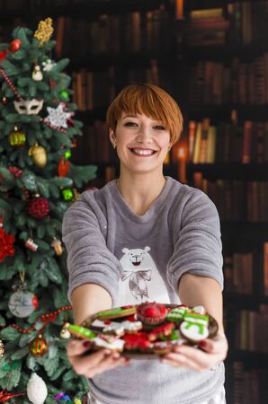 Portrait of beautiful redhead woman holding plate with christmas cakes before dressed up treeの写真素材
