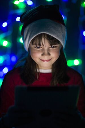 Young smiling girl in santa's hat holding tablet pc before bokeh lights background in dark roomの写真素材