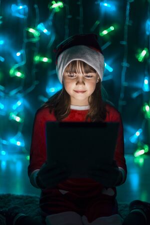 Young smiling girl in santa's hat holding tablet pc before bokeh lights background in dark roomの写真素材