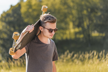 Cute young hipster boy wears sunglasses holding longboard on his shoulder