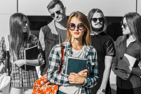 Cheerful blonde girl with red backpack in sunglasses holds book before modern buildingの写真素材