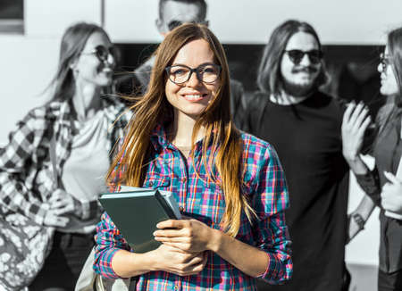 Cheerful brunette girl with white backpack holds books, glasses and headphones before modern buildingの写真素材