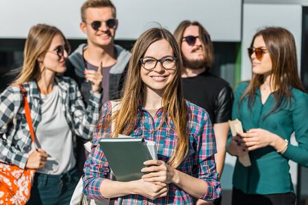 Cheerful brunette girl with white backpack holds books, glasses and headphones before modern buildingの写真素材