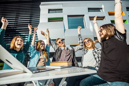 Teamwork cooperation emotions of group of students, happy successful emotions, table with laptop and books before modern building, sunny dayの写真素材