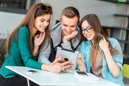 Group of students wear glasses have work conversation outdoor cafe, table with tablet computer before modern building wall, sunny dayの写真素材