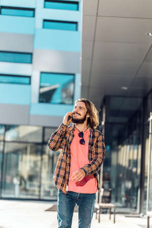 Happy smiling young man walking and talk phone before modern buildingの写真素材