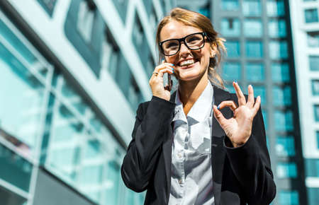 Young blonde caucasian businesswoman wears glasses, white blouse and a black jacket is talking phone shows OK sign before the modern corporation building in the backgroundの写真素材