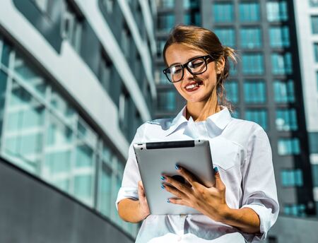 Young blonde caucasian businesswoman wears glasses and white blouse use tablet pc before the modern corporation building in the backgroundの写真素材