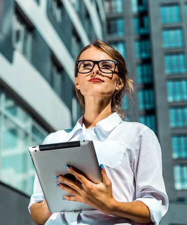 Young blonde caucasian businesswoman wears glasses and white blouse use tablet pc before the modern corporation building in the backgroundの写真素材