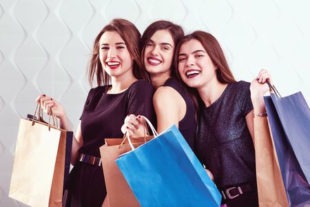 Friends shopaholic smiling girls holding colorful shopping bags before white indoor backgroundの写真素材