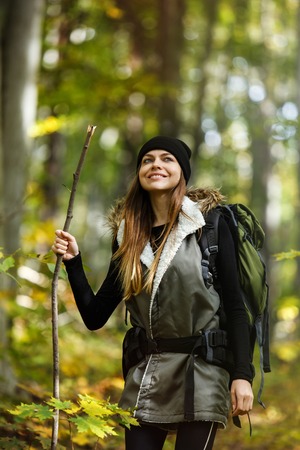 Cheerful brunette tourist girl wears black cap and backpacked have walk through forest holding stick, autumn tourism conceptの写真素材