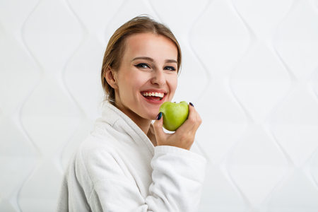 Cheerful woman in the white bathrobe enjoys a green apple, indoor shot in the white backgroundの写真素材