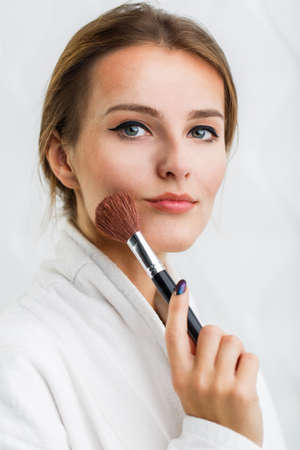 Lovely girl in the white bathrobe using makeup brush in the white backgroundの写真素材