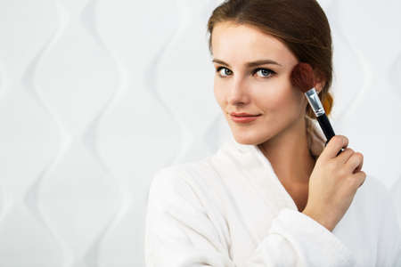 Lovely girl in the white bathrobe using makeup brush in the white background