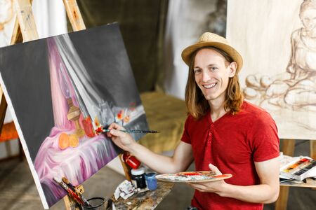Long hair caucasian male artist, in a light brown hat, smiling during painting in the studioの写真素材