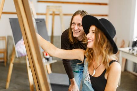 Male artist, in casual torn jeans, consulting his female colleague, in balck hat and denim dangarees, while drawingの写真素材