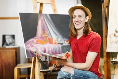 Long hair caucasian male artist, in a light brown hat, smiling during painting in the studioの写真素材