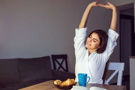Beautiful caucasian girl in a white bathrobe enjoys morning coffee and croissant, time in the kitchenの写真素材