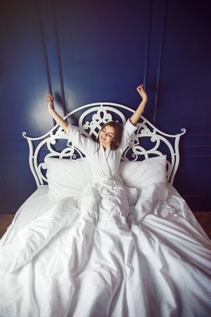 Beautiful caucasian girl, in a white bathrobe, enjoys morning in the bedroom before blue wallの写真素材